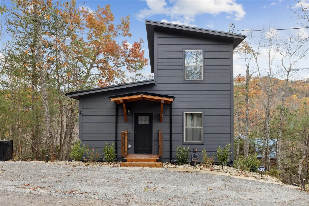 The image showcases the striking exterior of a brand-new, modern cabin nestled in the woods of Sevierville, Tennessee. A spacious wooden porch overlooks a thoughtfully landscaped yard, perfect for gatherings, grilling, or simply soaking in the serene mountain atmosphere. The inviting setting is ideal for relaxing, watching sunsets, and enjoying nature just minutes from the Smoky Mountains.