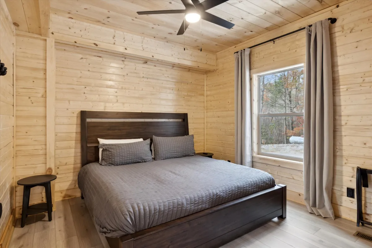 This image features a serene bedroom with a neatly made bed, a large window allowing soft natural light to enter, and a nightstand beside the bed. The space is uncluttered and emphasizes calmness, ideal for rest and relaxation.