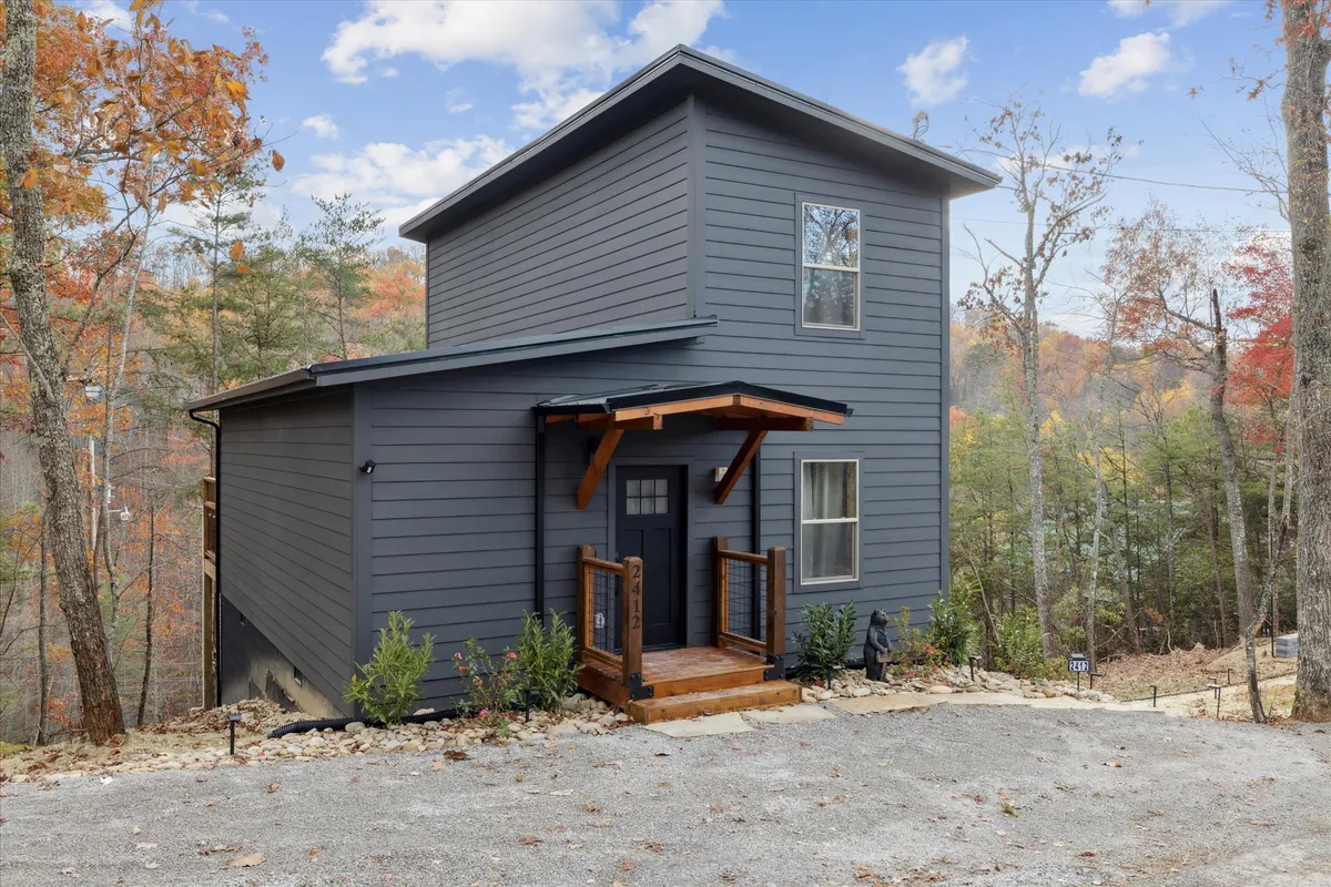 A welcoming exterior view of the Black Bear Cabin, showcasing its modern architecture, inviting wooden porch, and rich surrounding greenery. The setting is framed by mature trees, hinting at the natural seclusion and opportunities for outdoor gatherings, nature walks, or simply relaxing outside.