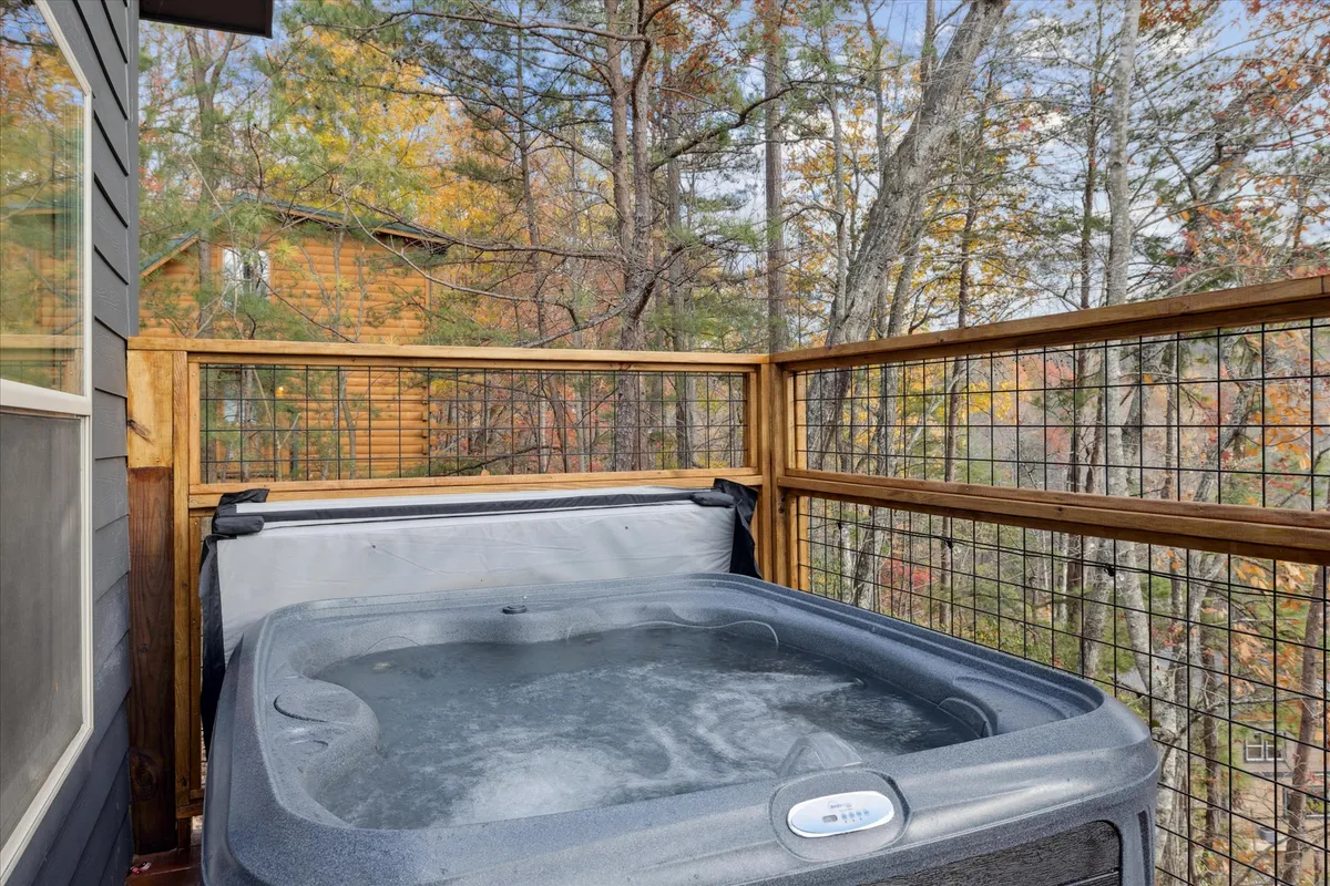 This image showcases a tranquil hot tub set on a wooden deck, surrounded by lush trees and protected by a rustic railing. The scene exudes relaxation, with the bubbling water inviting guests to unwind while soaking in the beauty of the natural landscape.