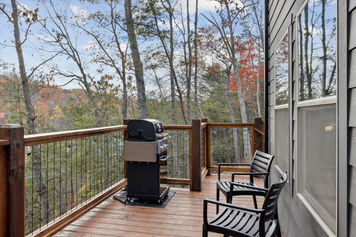 A cozy outdoor deck features a modern grill, comfortable deck chairs, and a wooden railing against a scenic backdrop of trees. The setting is ideal for barbecuing, enjoying meals outdoors, or simply relaxing in nature.