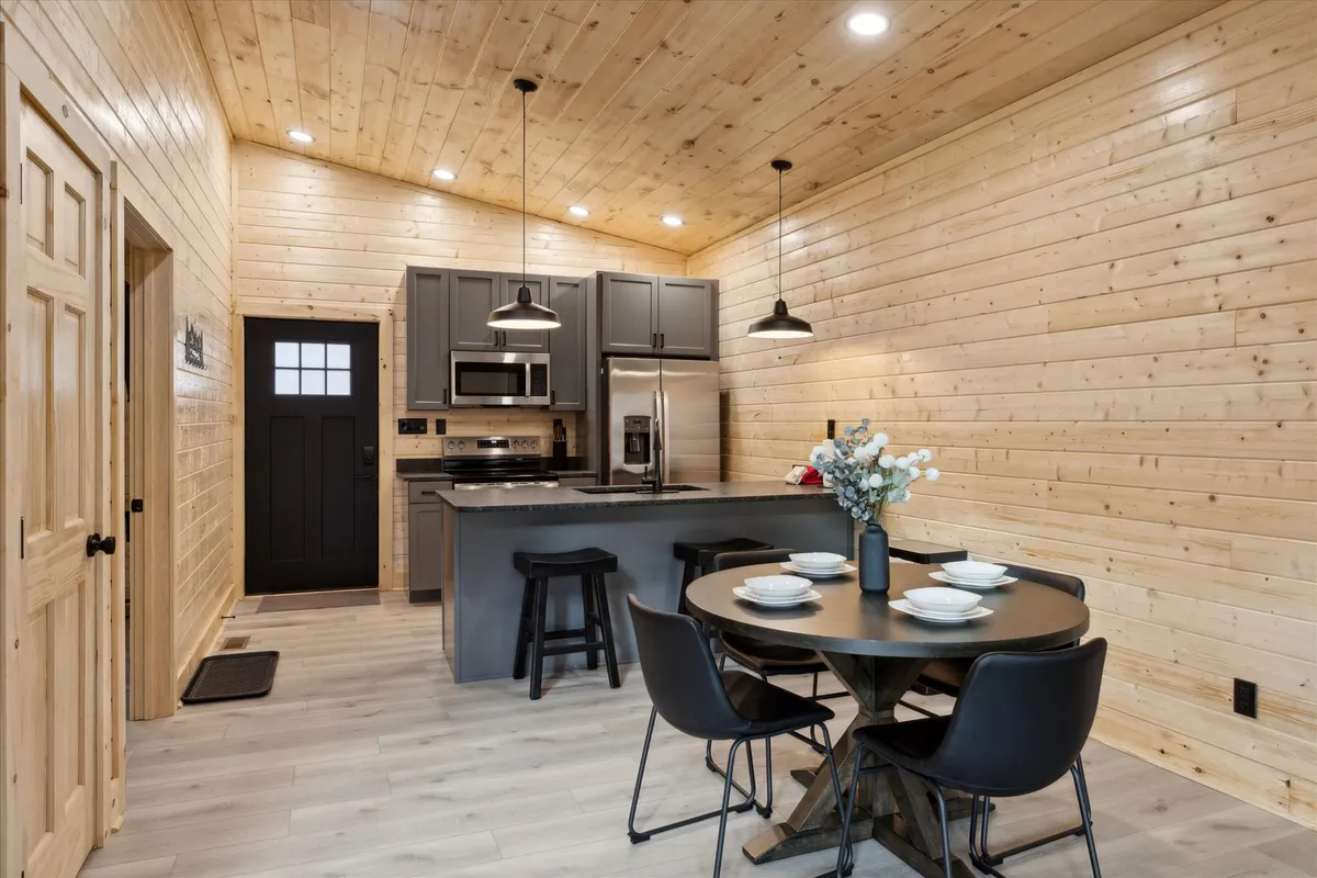 A cozy, welcoming kitchen and dining area with a central kitchen island, ample wooden cabinetry, and a dining table set for meals. Warm wood walls and natural light create a homey atmosphere, inviting guests to cook, dine, and gather together.