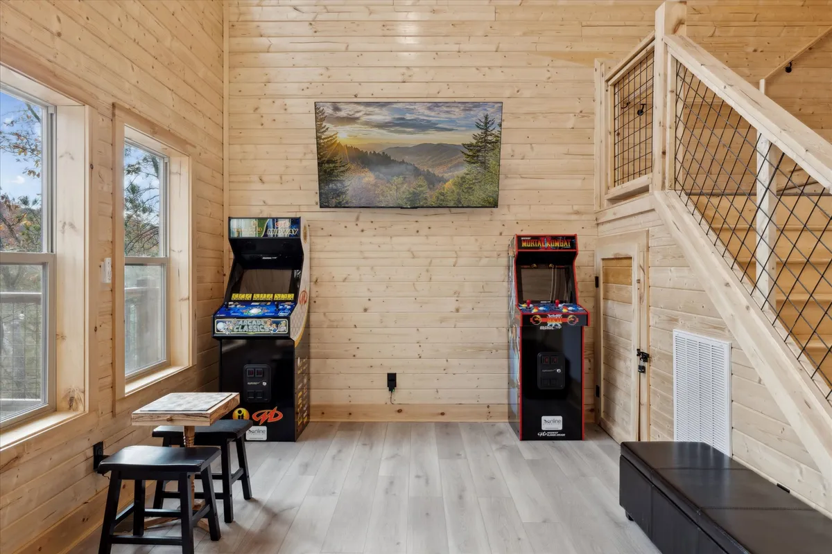 A vibrant interior game room featuring multiple arcade machines against a wall adorned with landscape artwork. Stools are positioned for easy gameplay, and a staircase hints at the open flow of the space. The room invites fun and friendly competition in a modern rustic setting.